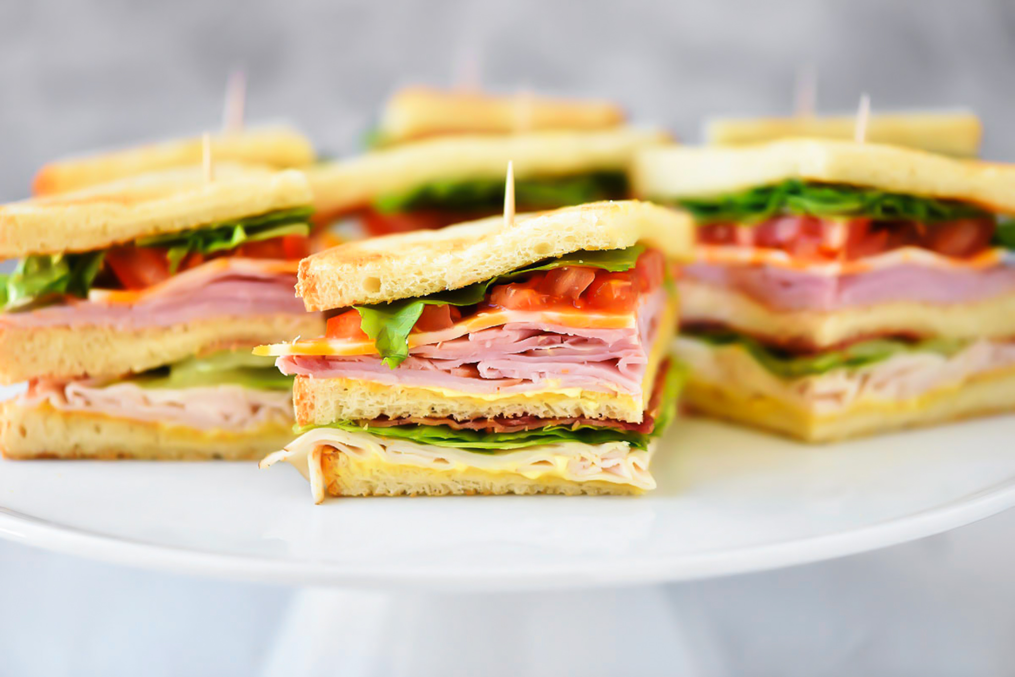 Club Sandwiches are stacked with ham, turkey, cheese, lettuce and tomato with a mustard and mayo mixture.