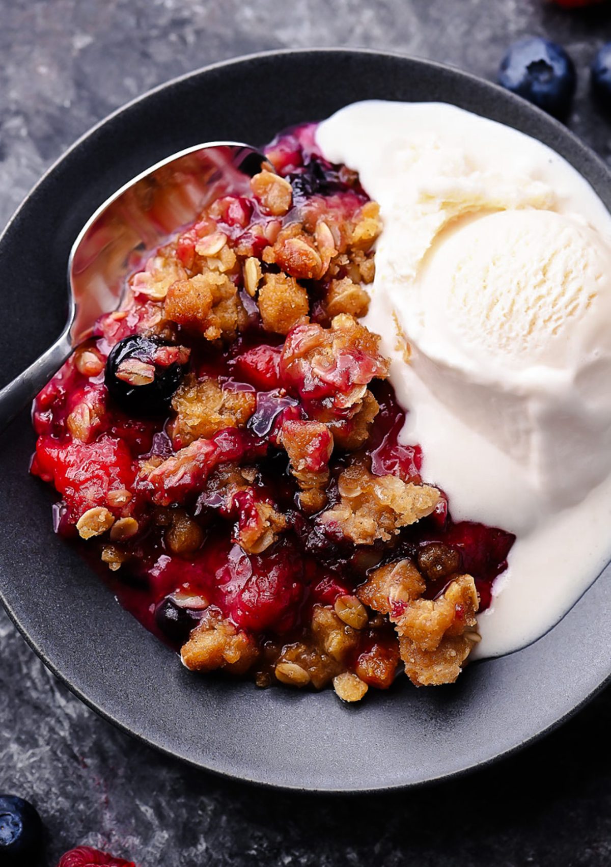 A warm berry crisp with bubbling mixed berries and a golden oat crumble topping, served in a white dish with a scoop of melting vanilla ice cream on top.